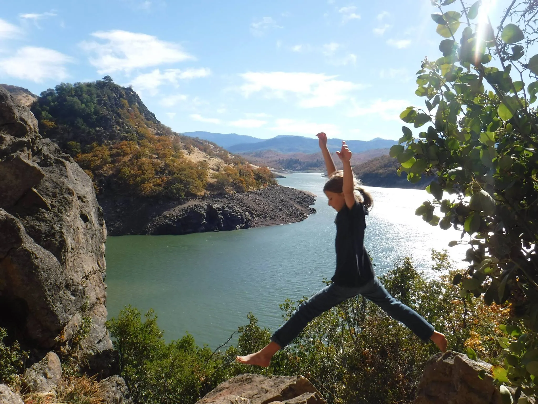 A participant standing with arms raised on rocks above a reservoir, oak-covered ridges behind