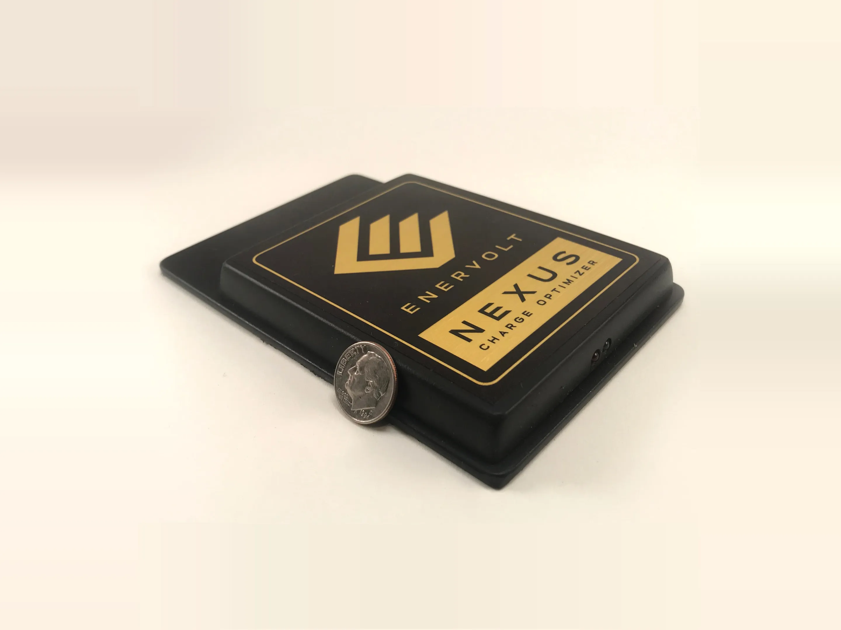 The Nexus Charge Optimizer unit photographed beside a dime for scale, its moulded case carrying the Enervolt mark and the Nexus Charge Optimizer label in gold