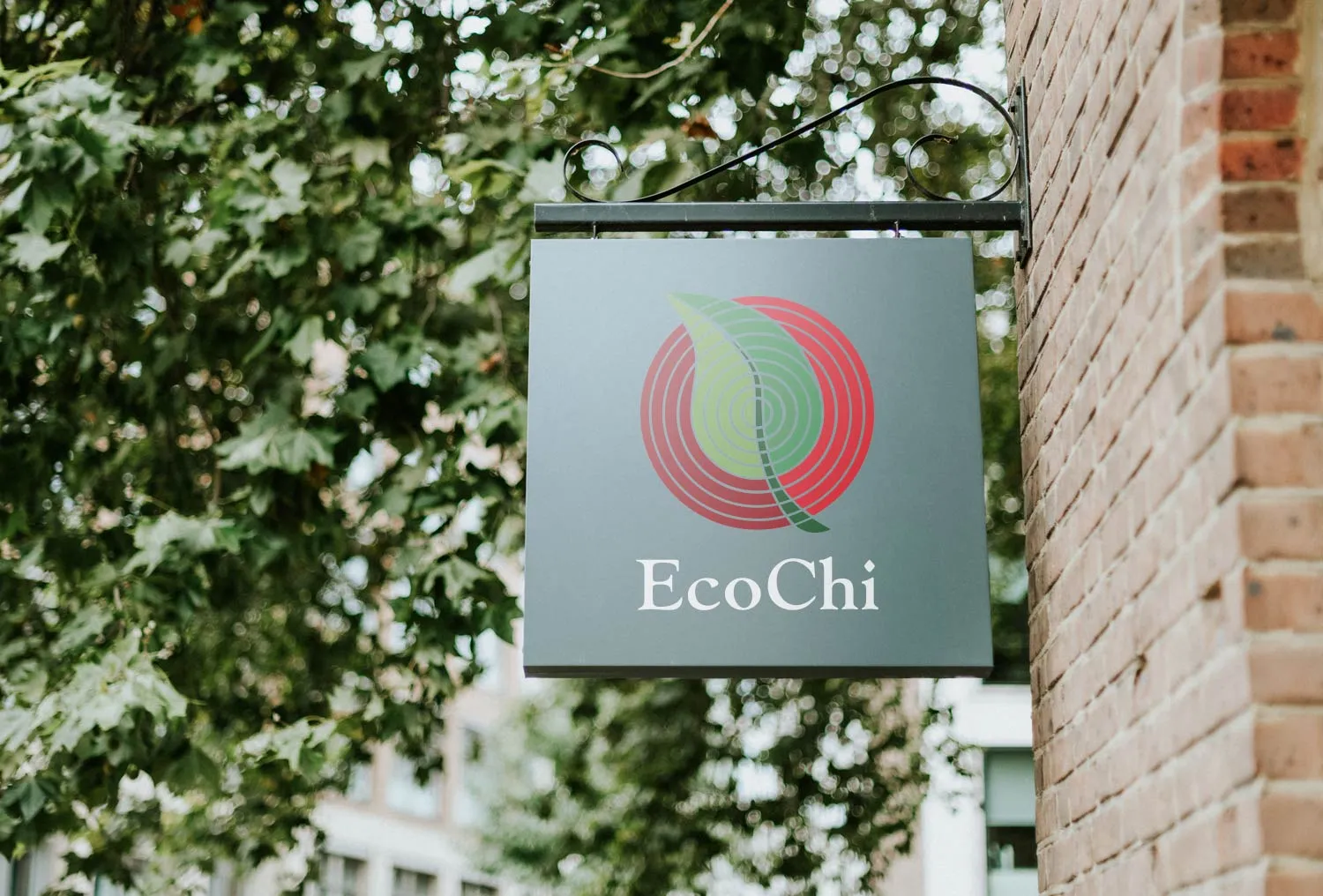 An EcoChi sign hanging from a bracket on a brick building, the mark and wordmark set on a slate ground against summer trees