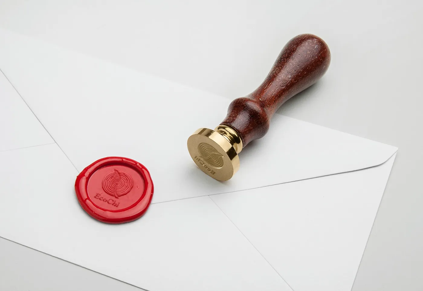 A brass EcoChi wax stamp resting on a white envelope beside a pressed red wax seal bearing the mark