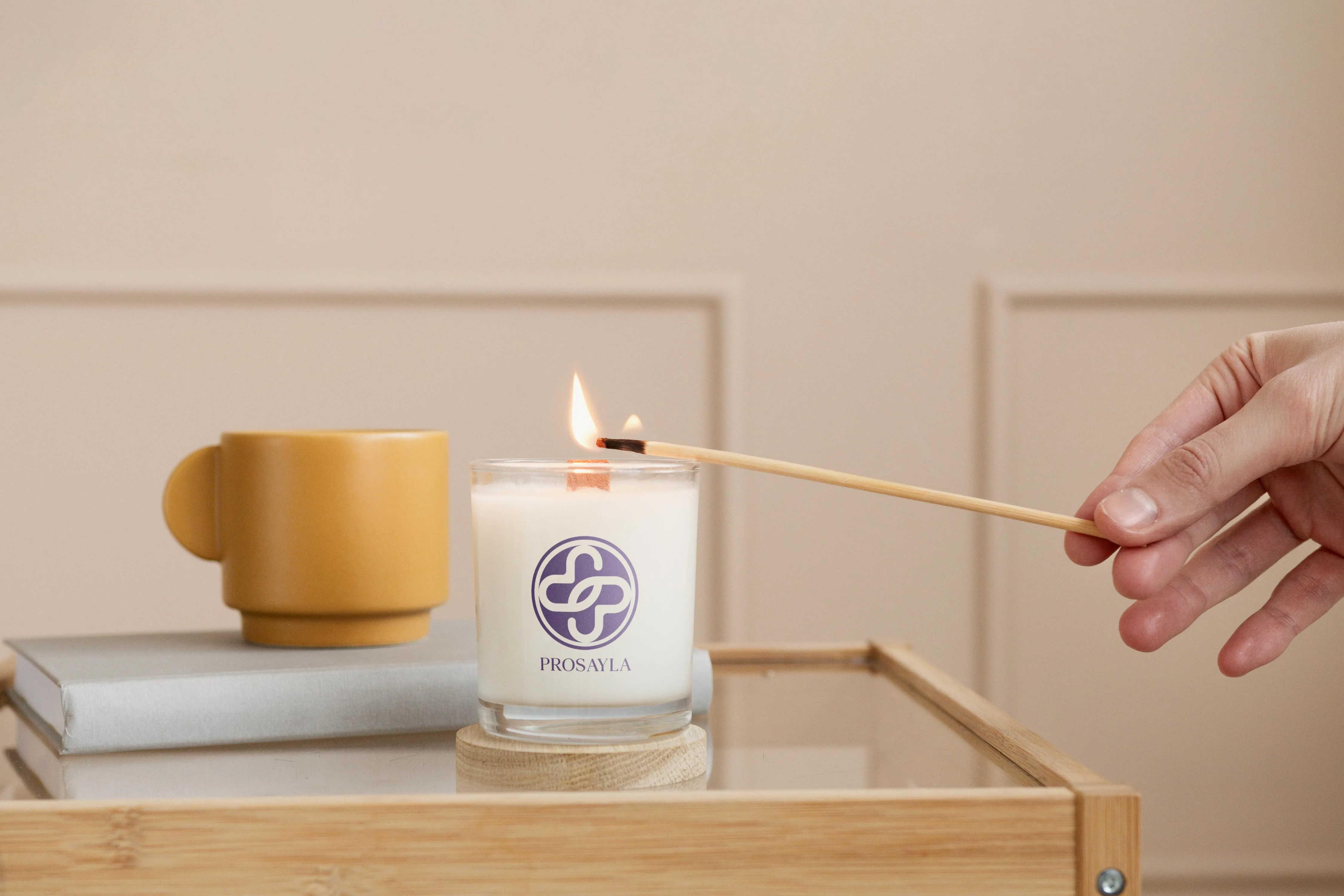 A Prosayla-branded candle being lit with a long match, the purple interlocking mark printed on the glass
