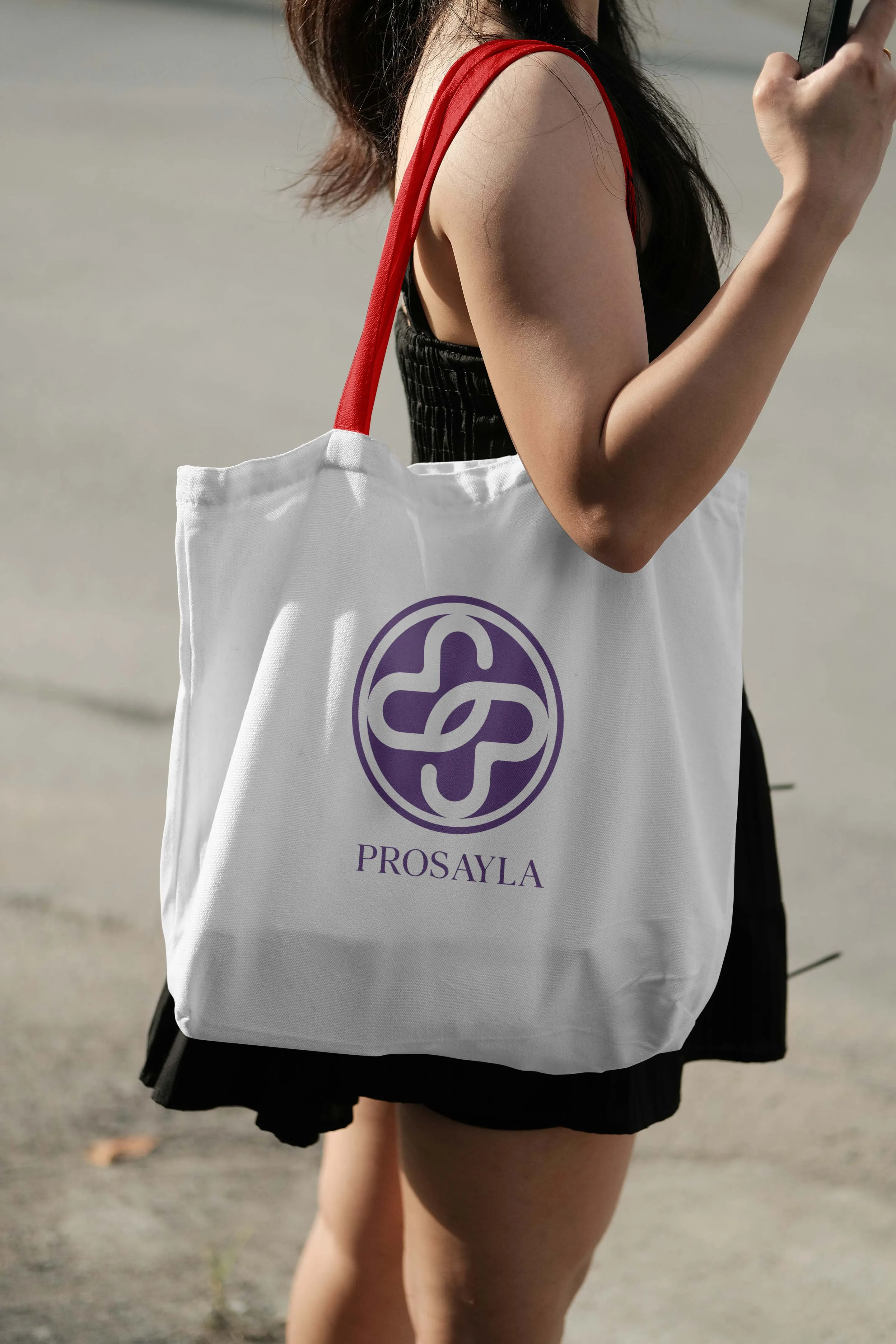 A white canvas tote carried over a shoulder, printed with the purple Prosayla mark and wordmark