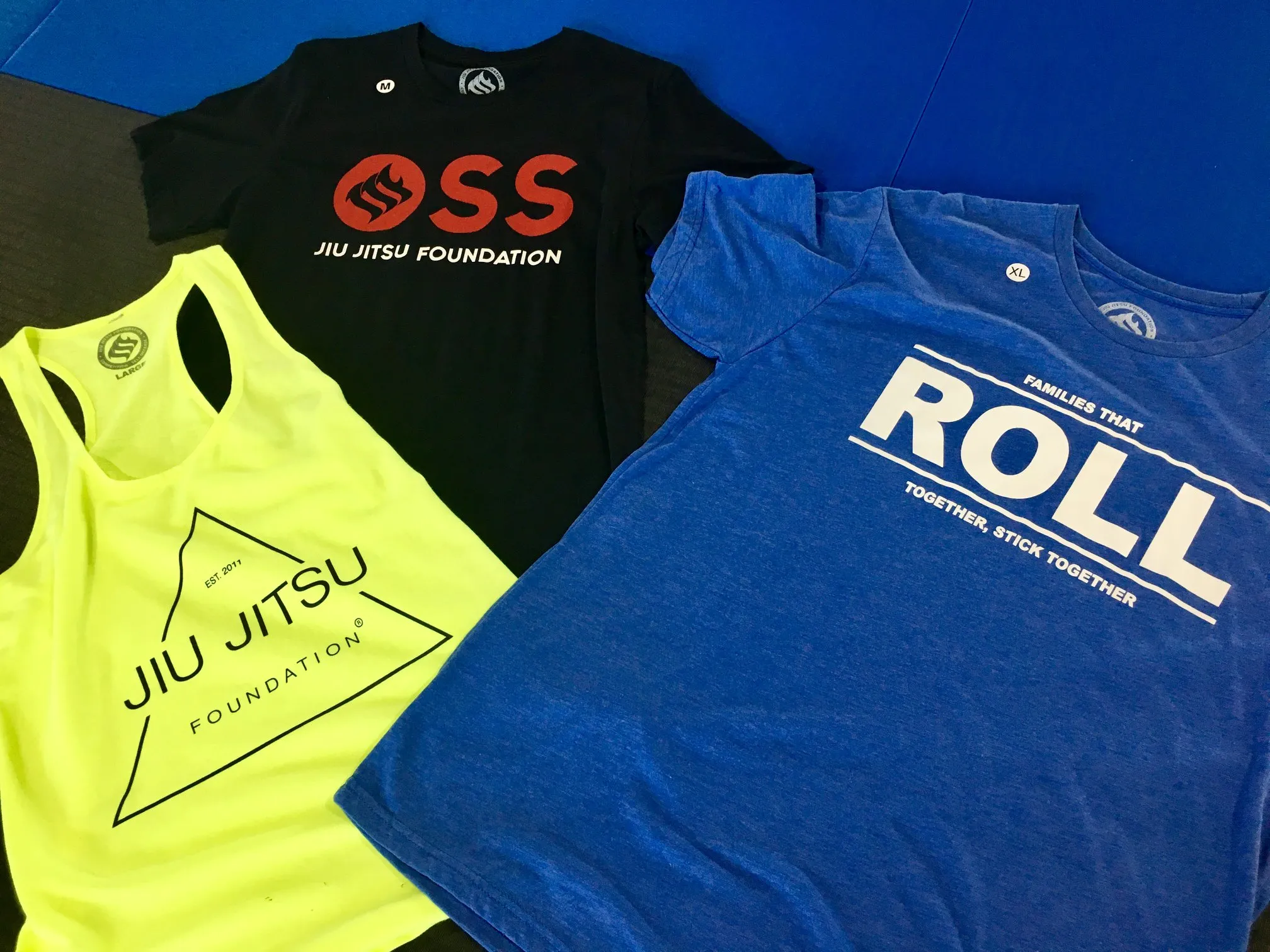 Academy apparel laid out: a black OSS tee, a neon training tank, and a blue ROLL shirt