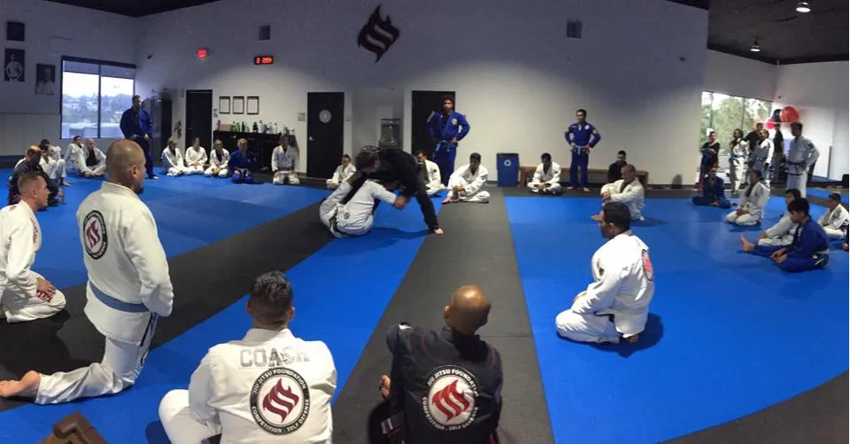 A full class seated along the mats, coaching staff in patched gis at the front