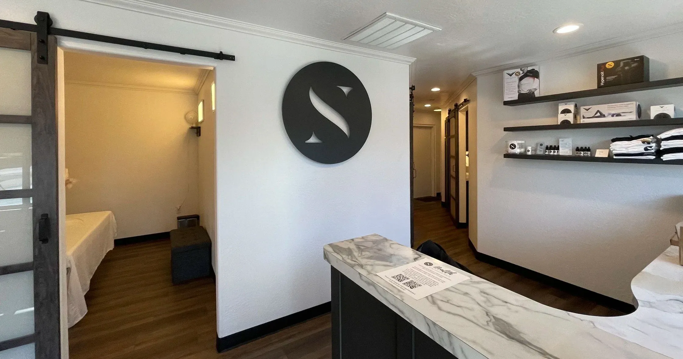 The Nova Sana reception: the circular S mark mounted on the wall, a marble desk, a retail shelf, and a treatment room through an open sliding door