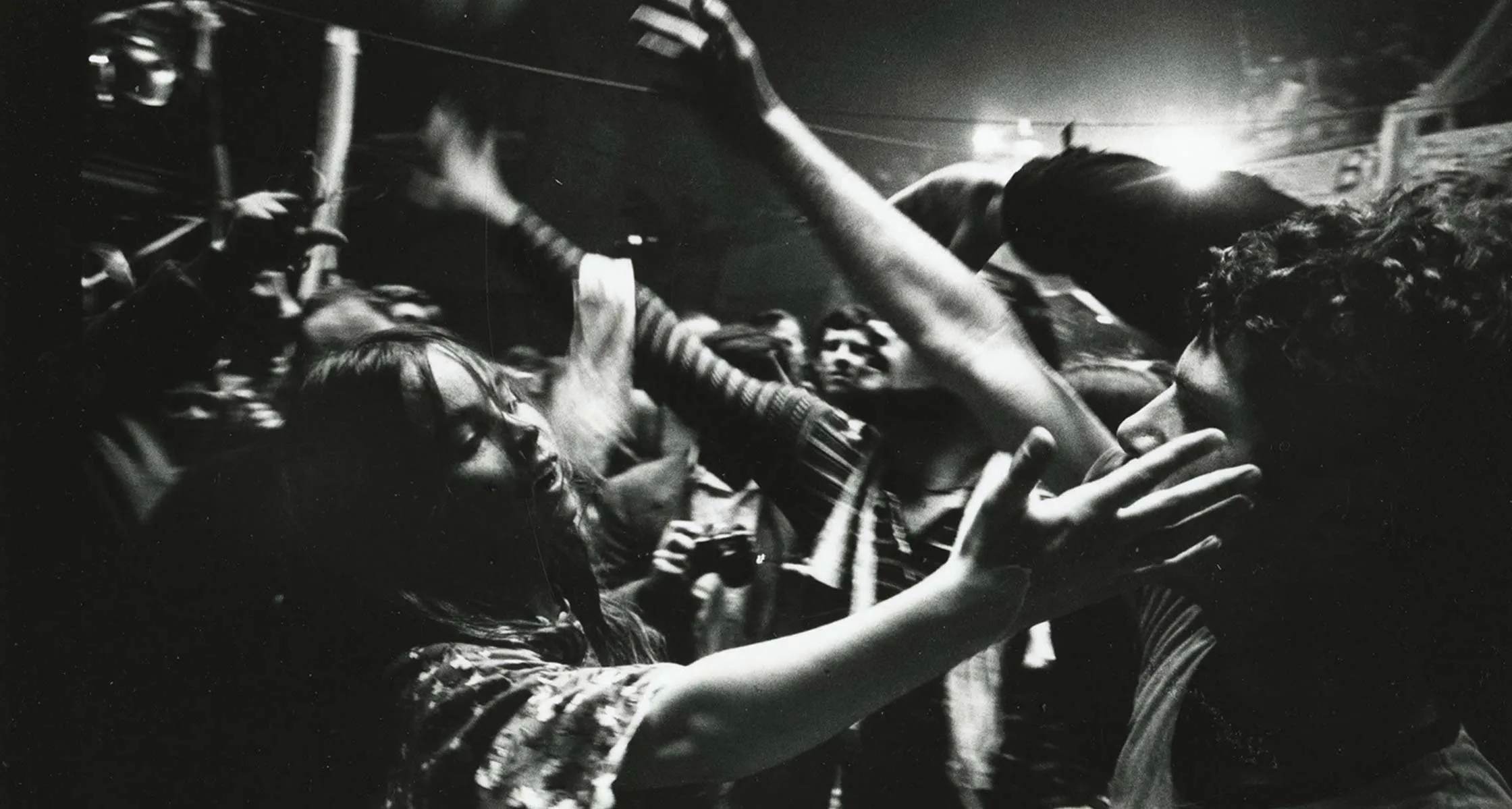 A black and white photograph of a crowd dancing at a live show, arms raised in motion