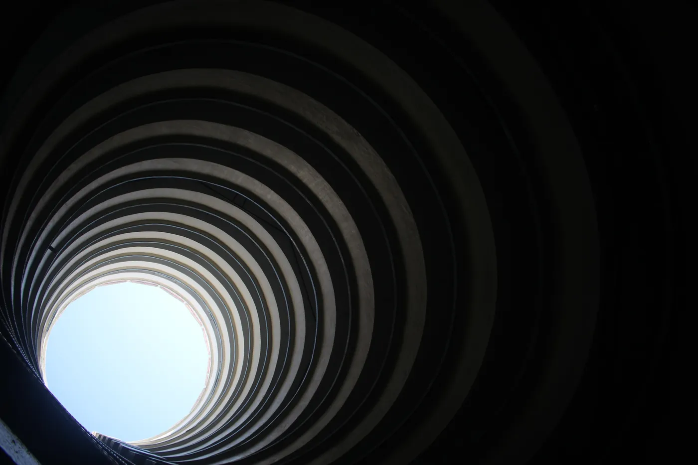 A view straight up through the concentric rings of a spiral concrete structure toward a small circle of pale sky