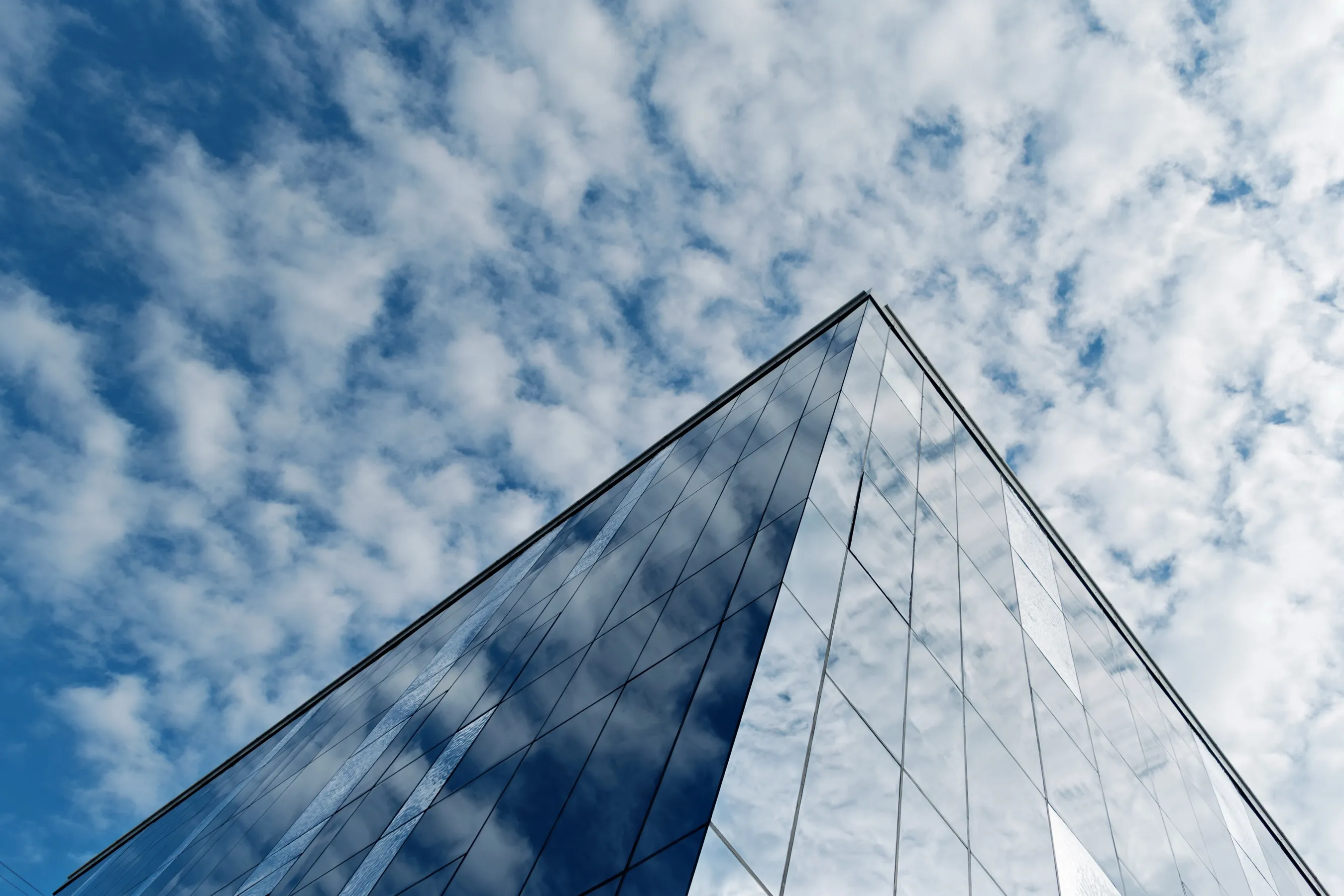 The corner of a glass building where two mirrored facades reflect the same cloudy sky differently